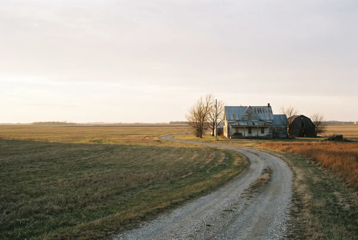 A farmhouse with a metal roof at the end of a gravel road through open farmland, under a wide pale sky.